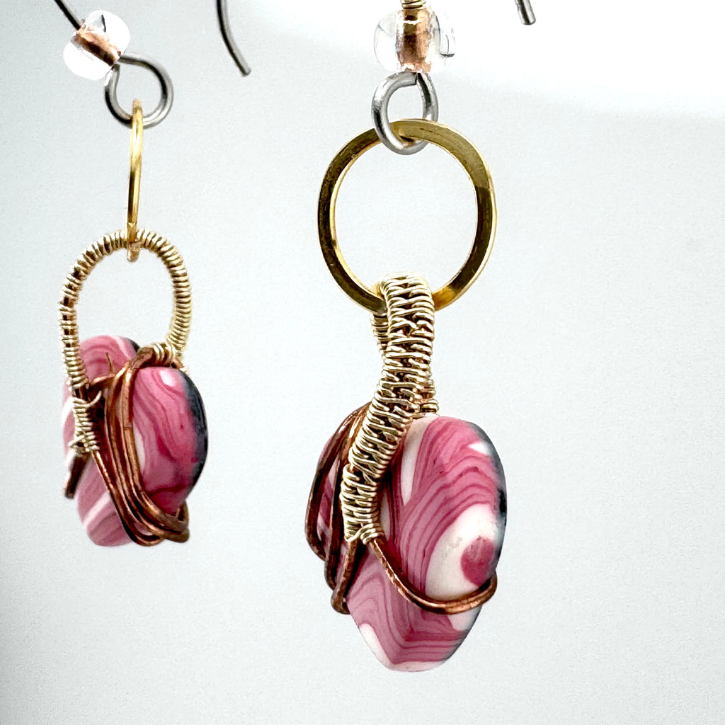 side view of a pair of handmade dangle earrings featuring marbled pink and white clay beads wrapped in textured gold and copper wire, suspended from silver-toned fishhook ear wires.