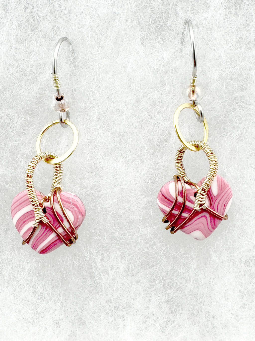 A pair of handmade dangle earrings featuring pink and white marbled heart-shaped clay beads wrapped in gold and rose gold wire, suspended from silver fishhook wires on a textured white background.