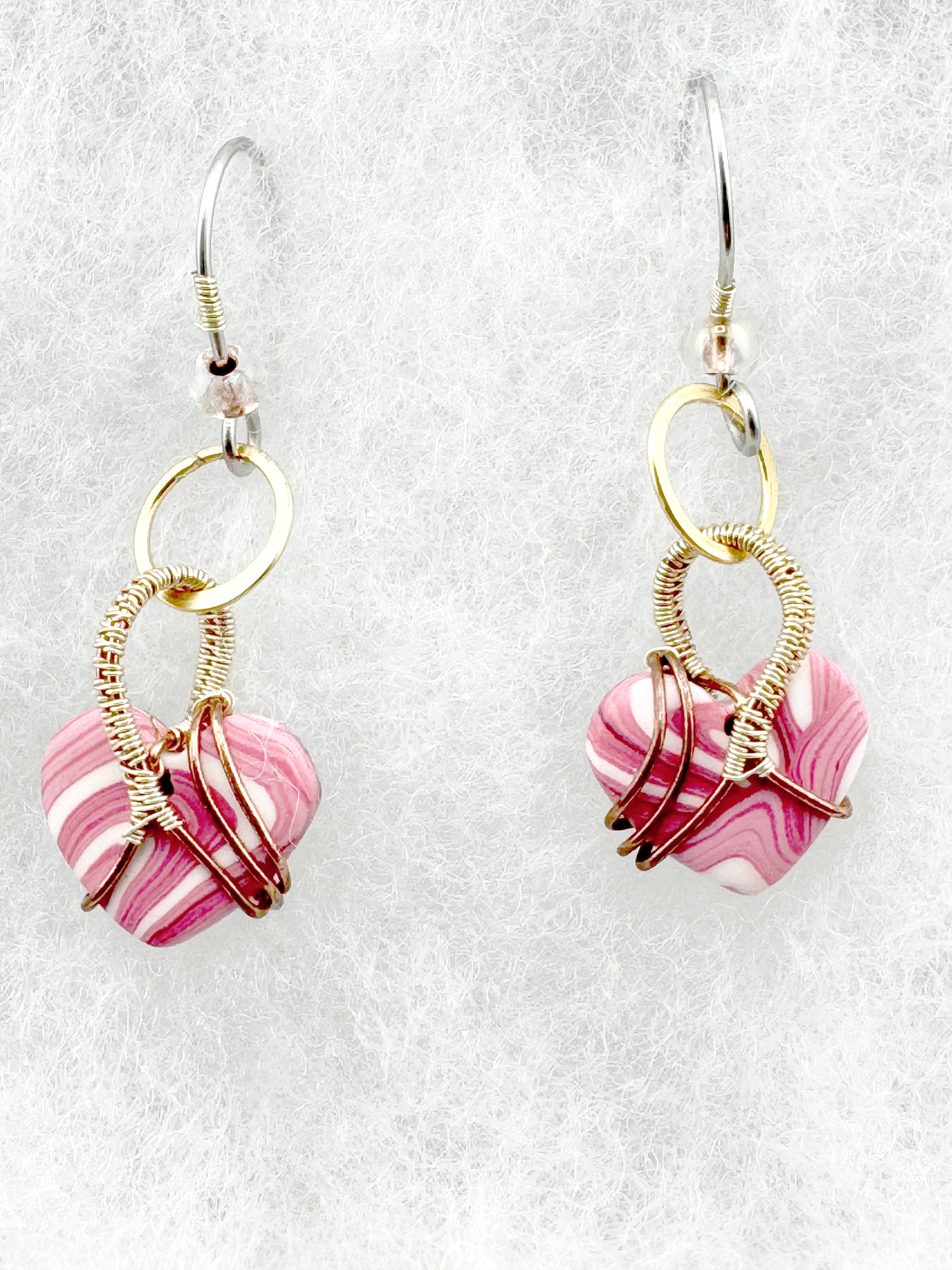 A pair of handmade dangle earrings featuring pink and white marbled heart-shaped clay beads wrapped in gold and rose gold wire, suspended from silver fishhook wires on a textured white background.
