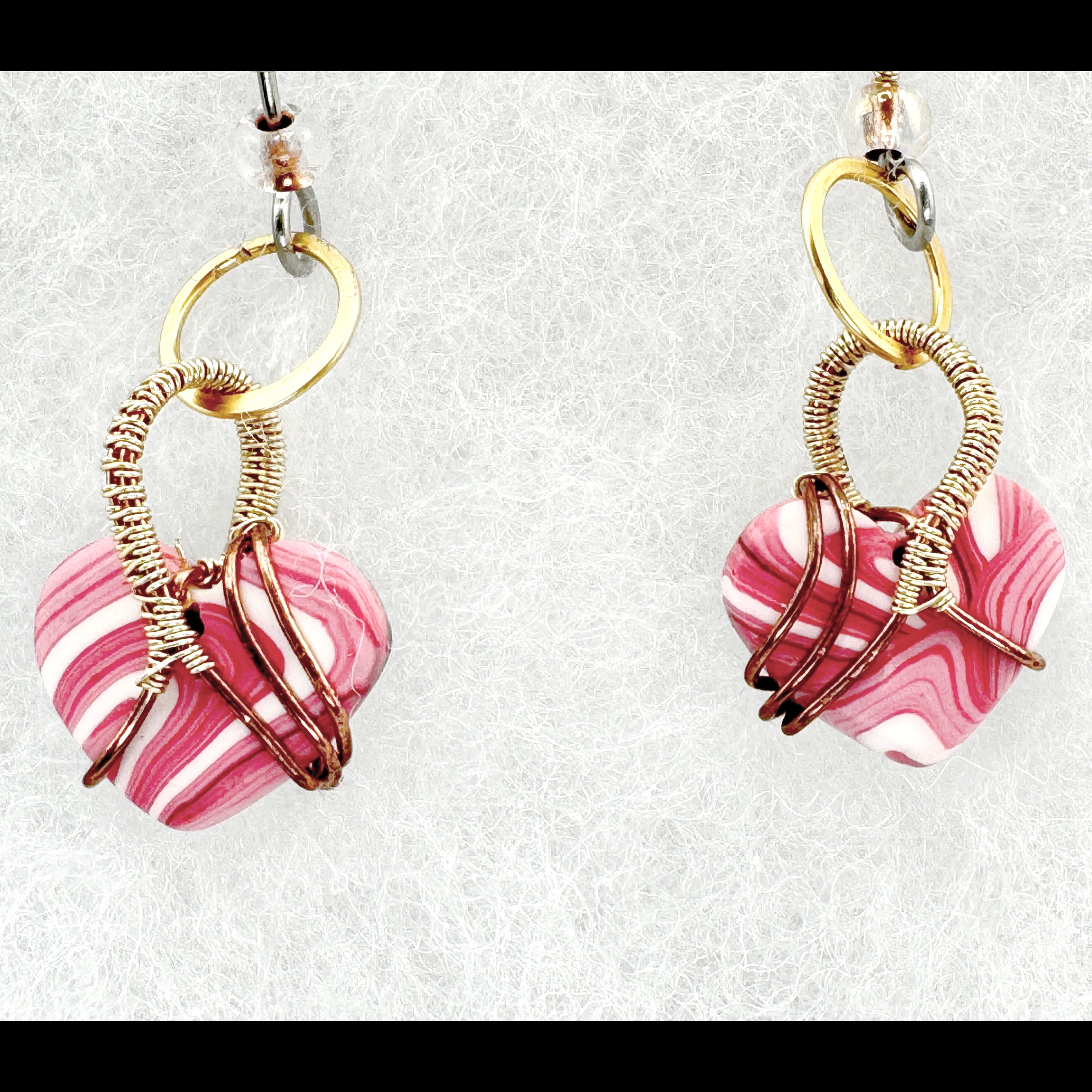 A pair of handmade heart-shaped dangle earrings featuring pink and white marbled clay beads wrapped in gold and copper wire, suspended from gold rings and silver ear wires on a textured white background.