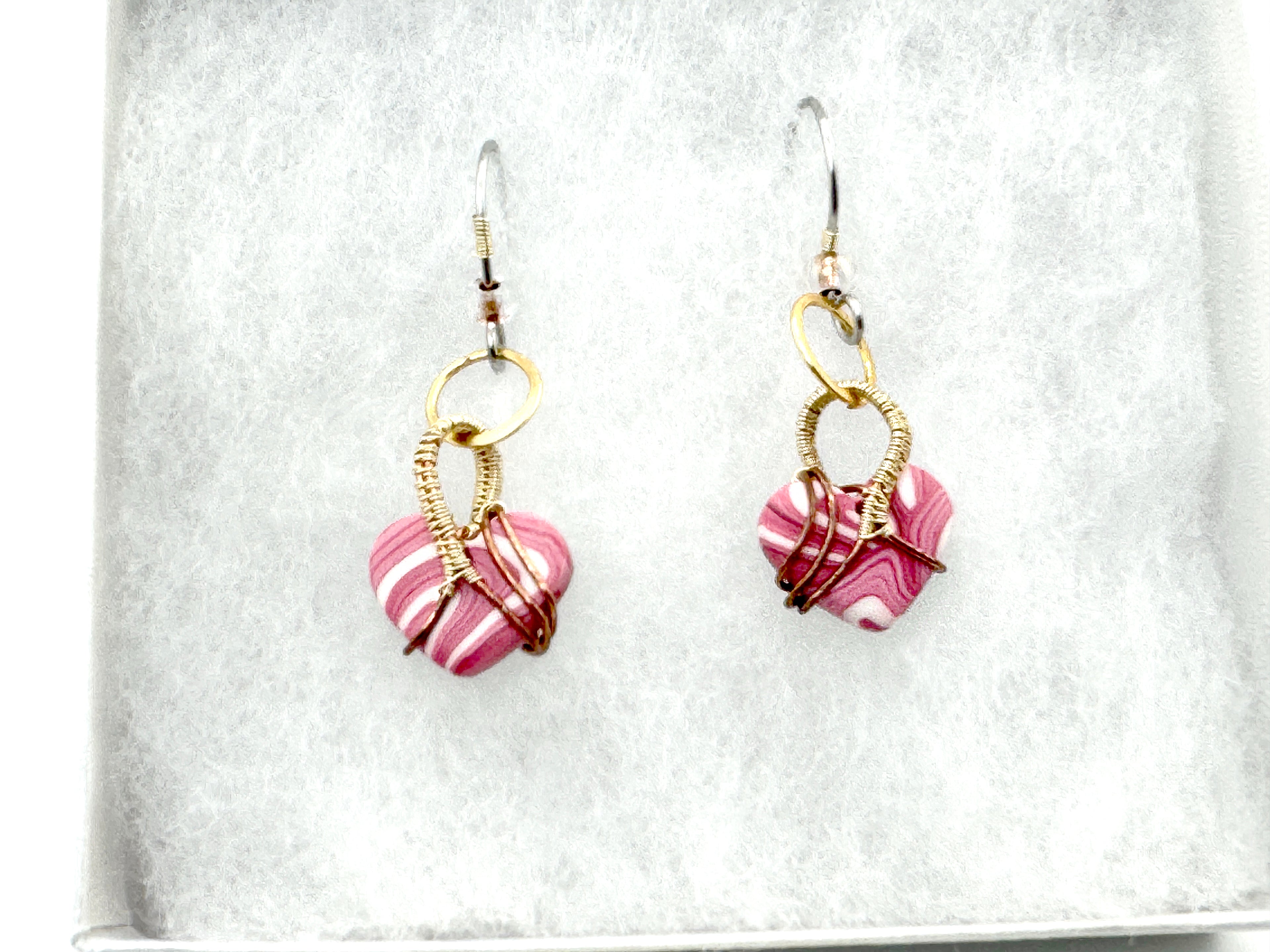 A pair of handmade dangle earrings with pink and white marbled clay heart  dangle earrings wrapped in gold wire, displayed on a textured white surface.