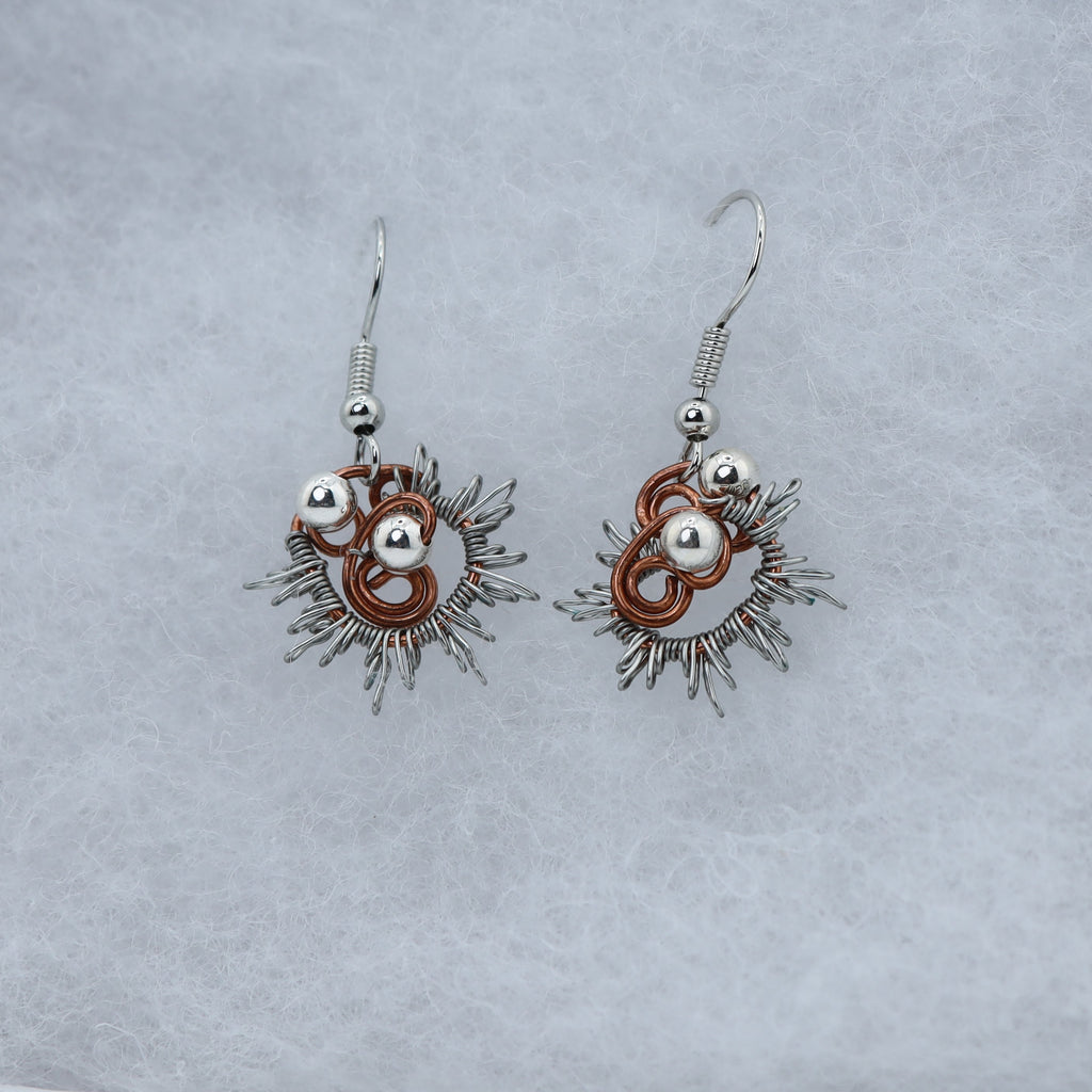 A pair of handmade dangle earrings with silver-toned metal, copper-toned rings, and silver beads, displayed on a textured light gray surface.