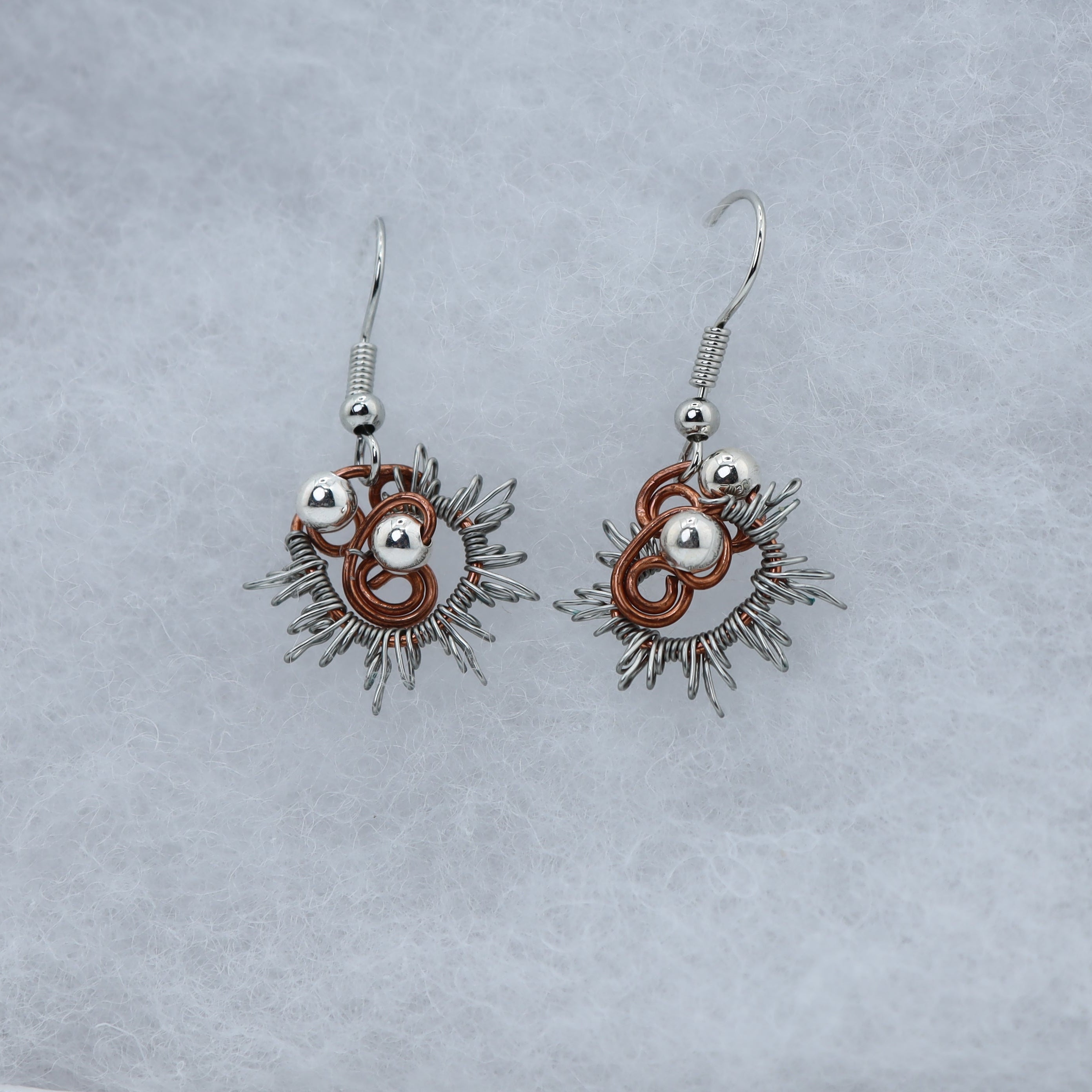 A pair of handmade dangle earrings with silver-toned metal, copper-toned rings, and silver beads, displayed on a textured light gray surface.