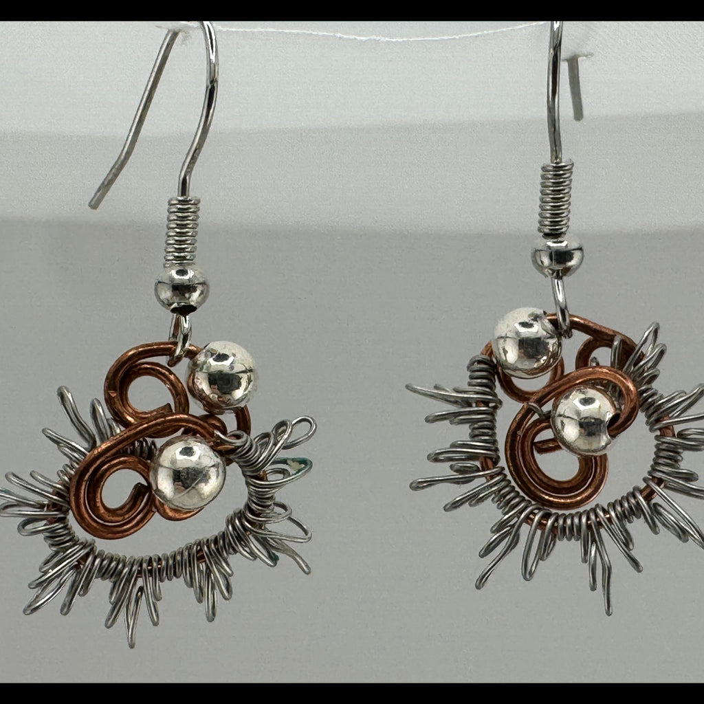A pair of handmade dangle earrings featuring silver-toned metal wire art with sunburst spikes, coiled copper wire accents, and small polished silver beads, hanging against a neutral light gray background.