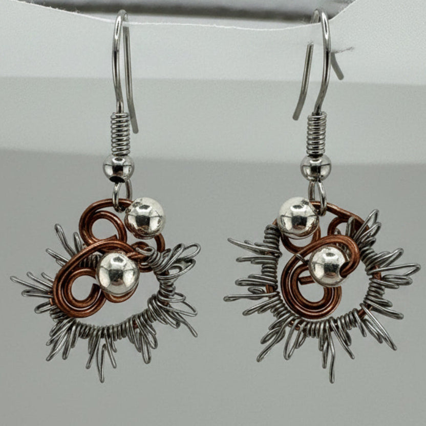 A pair of handmade dangle earrings featuring a copper and silver sunburst design with small silver beads, hanging from silver fishhook wires against a neutral gray background.