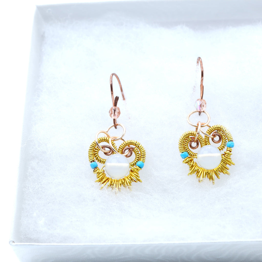 A pair of handmade dangle earrings displayed on a white textured insert inside a white gift box. The earrings feature a gold-toned sunburst design with turquoise blue accents and a white central stone, suspended from rose gold toned copper hooks.