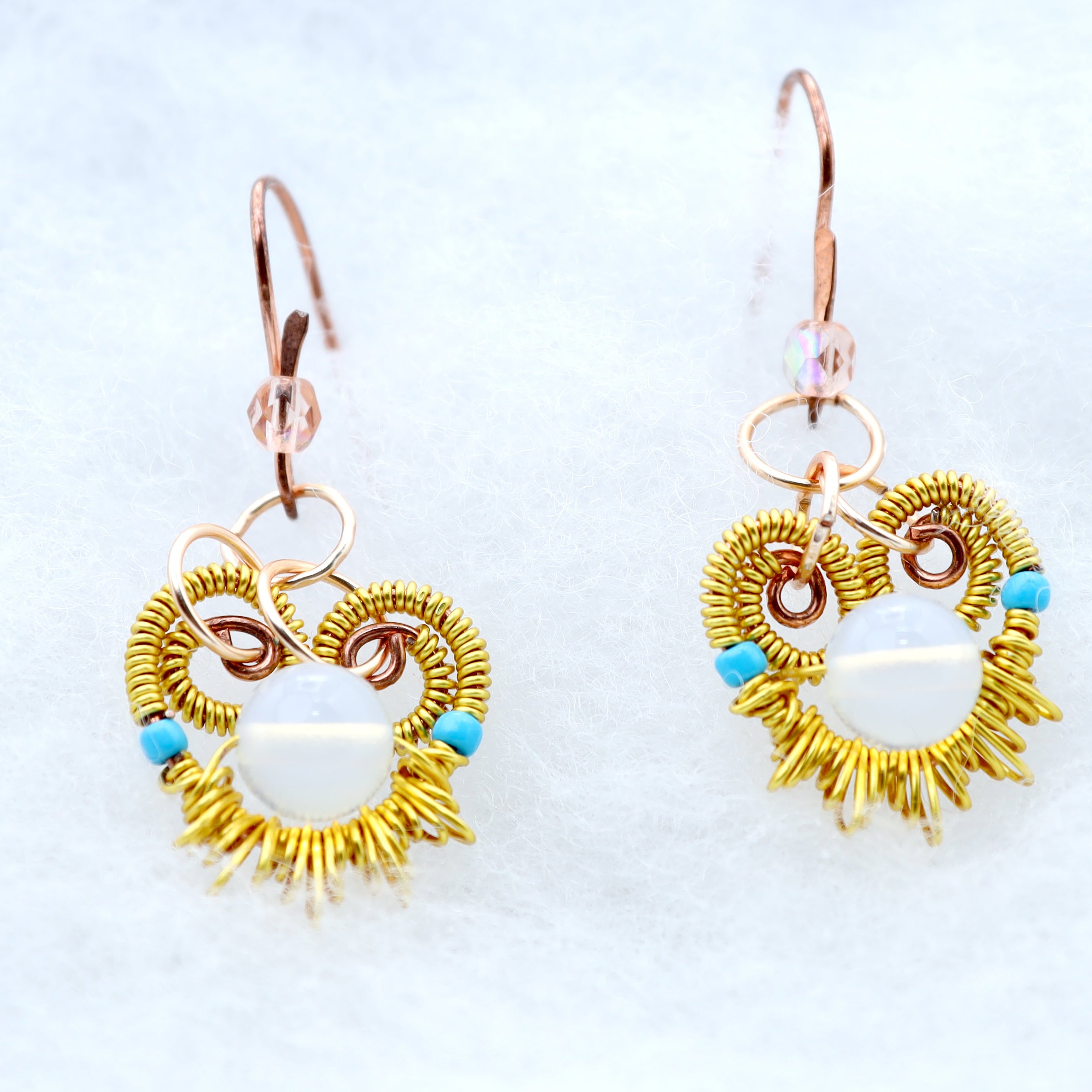 A pair of handmade dangle earrings featuring a gold sunburst wire design, rose gold toned copper hooks, and small turquoise blue beads, displayed on a white textured background.