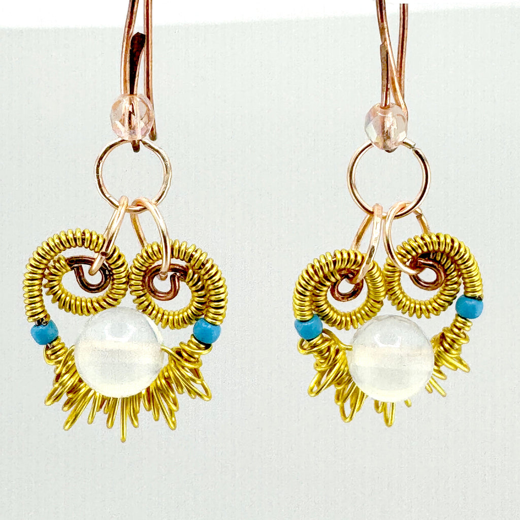 A pair of handmade dangle earrings featuring gold wire wrapped into a sunburst and owl-like circular patterns, with a large clear white oval bead in the center and small blue beads on either side. The earrings are suspended from rose gold toned copper hooks against a plain white background.