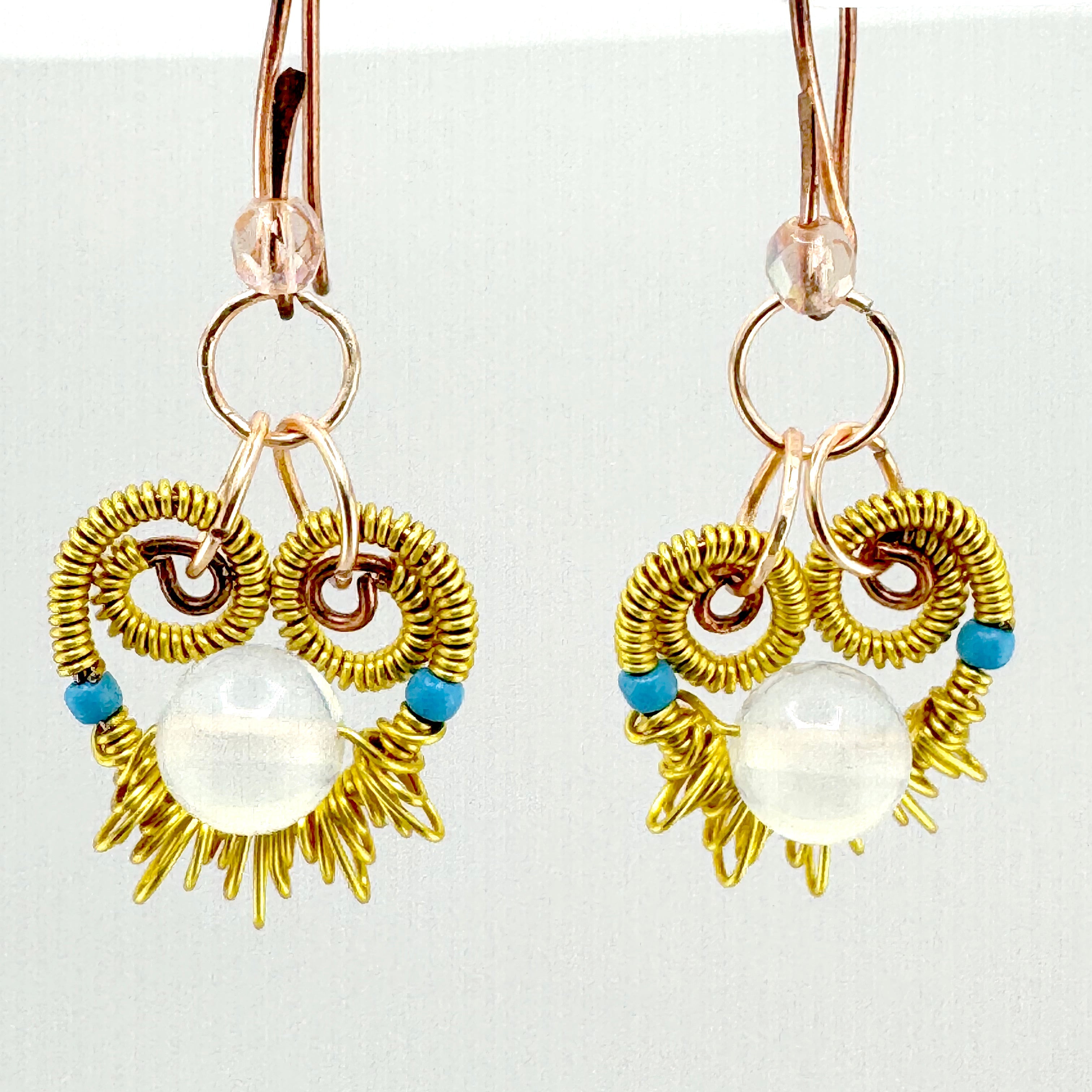 A pair of handmade dangle earrings featuring gold wire wrapped into a sunburst and owl-like circular patterns, with a large clear white oval bead in the center and small blue beads on either side. The earrings are suspended from rose gold toned copper hooks against a plain white background.