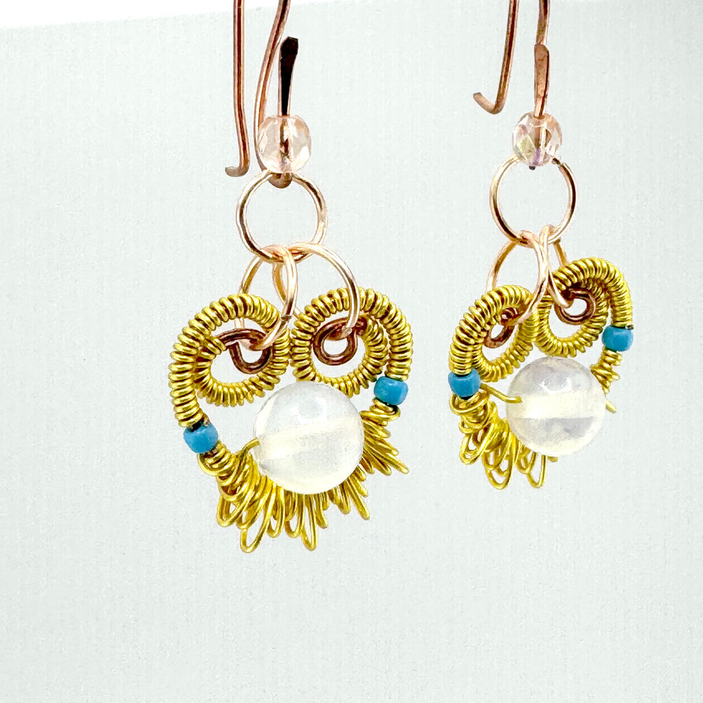 A pair of handmade dangle earrings featuring gold wire wrapped into a heart-like shape, accented with small blue beads and a large white round bead at the center, hanging from rose gold toned copper hooks against a plain white background.