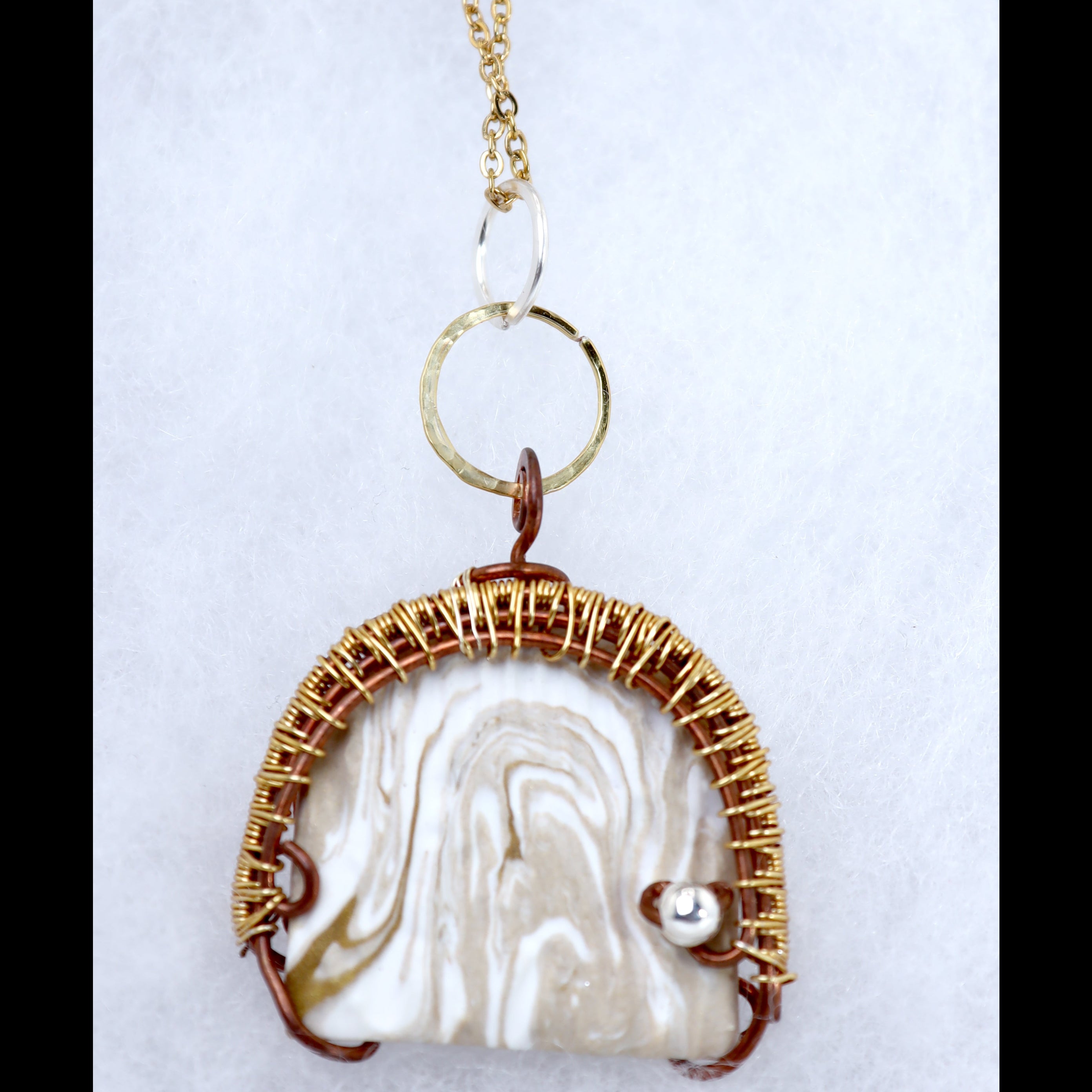 A handmade pendant necklace featuring a marbled cream and white clay piece wrapped in textured gold-toned wire, with a small silver accent and a gold ring connector on a white background.