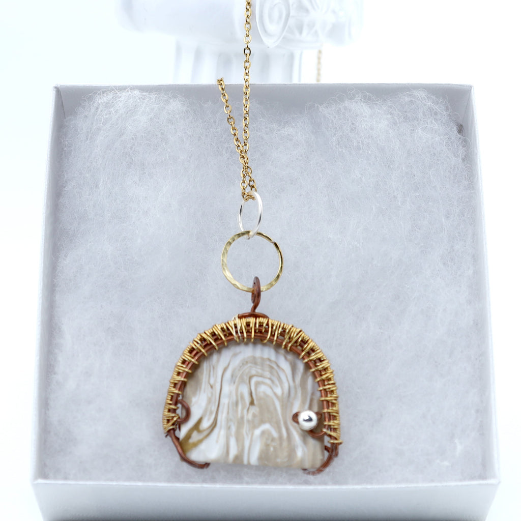 A gold-toned pendant necklace with a textured white and cream clay piece, displayed inside a light gray jewelry box.