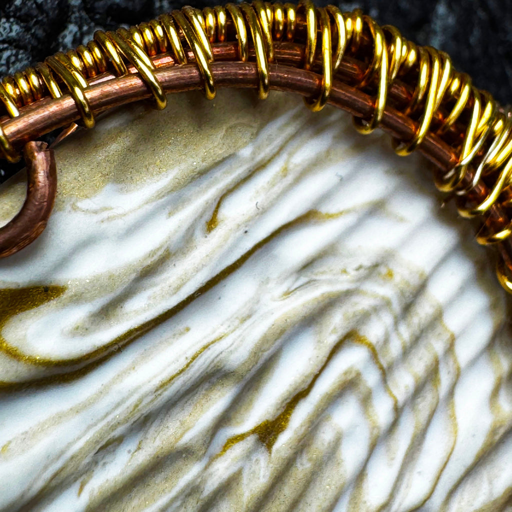 Close-up of a handmade wire-wrapped pendant featuring a textured, marbled white and gold stone in a copper and gold wire setting.