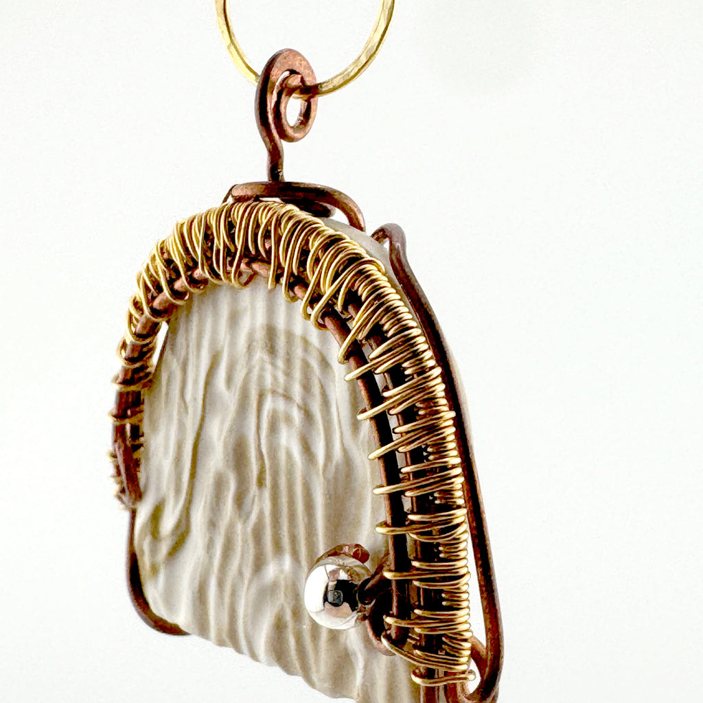 A handmade pendant featuring a textured cream-colored stone wrapped in copper and gold wire, with a small silver bead accent, shown against a plain white background.