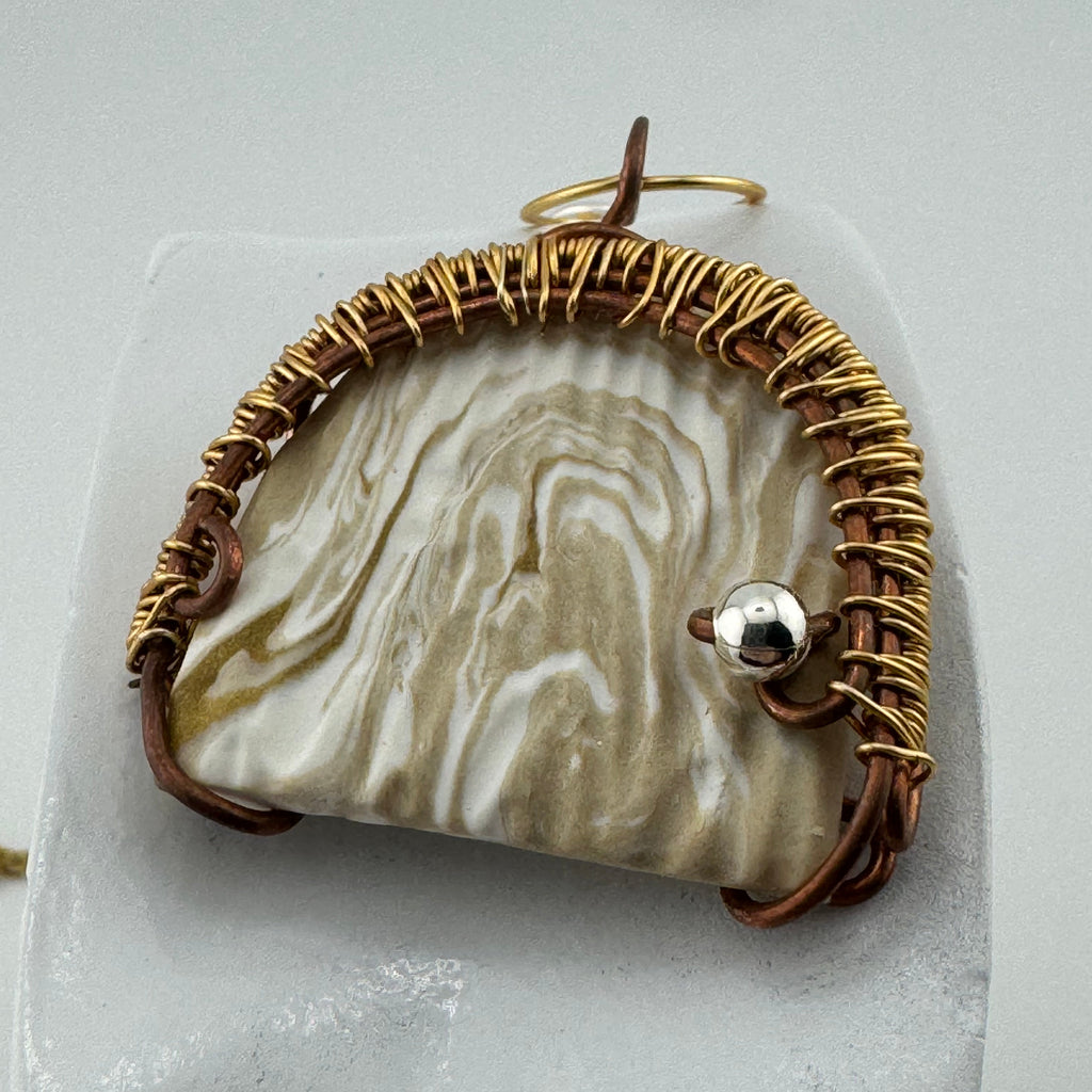 A handmade pendant featuring a  clay piece with natural beige and white marbled patterns, wrapped in textured gold wire and brown cord, with a small silver bead accent.