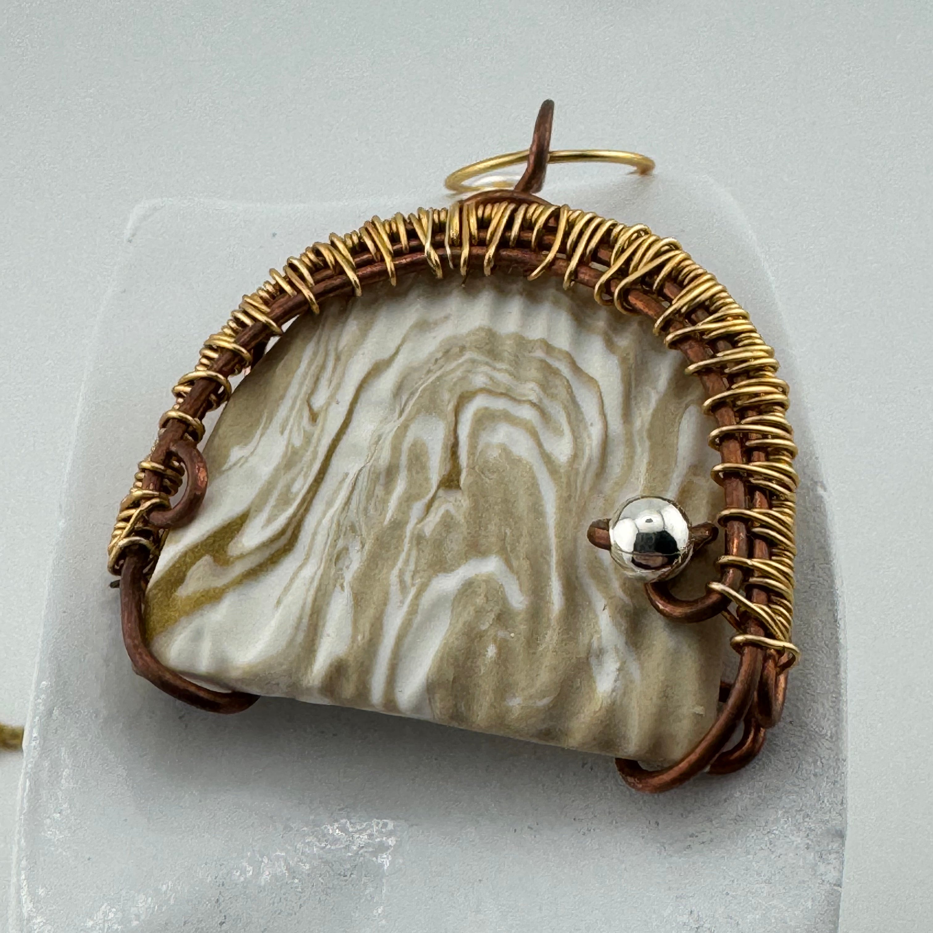 A handmade pendant featuring a  clay piece with natural beige and white marbled patterns, wrapped in textured gold wire and brown cord, with a small silver bead accent.