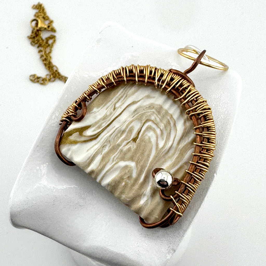 A handmade pendant featuring a cream and beige marbled stone set in a gold-toned bezel, wrapped in copper wire with a small silver bead displayed on a white stand with a gold chain.