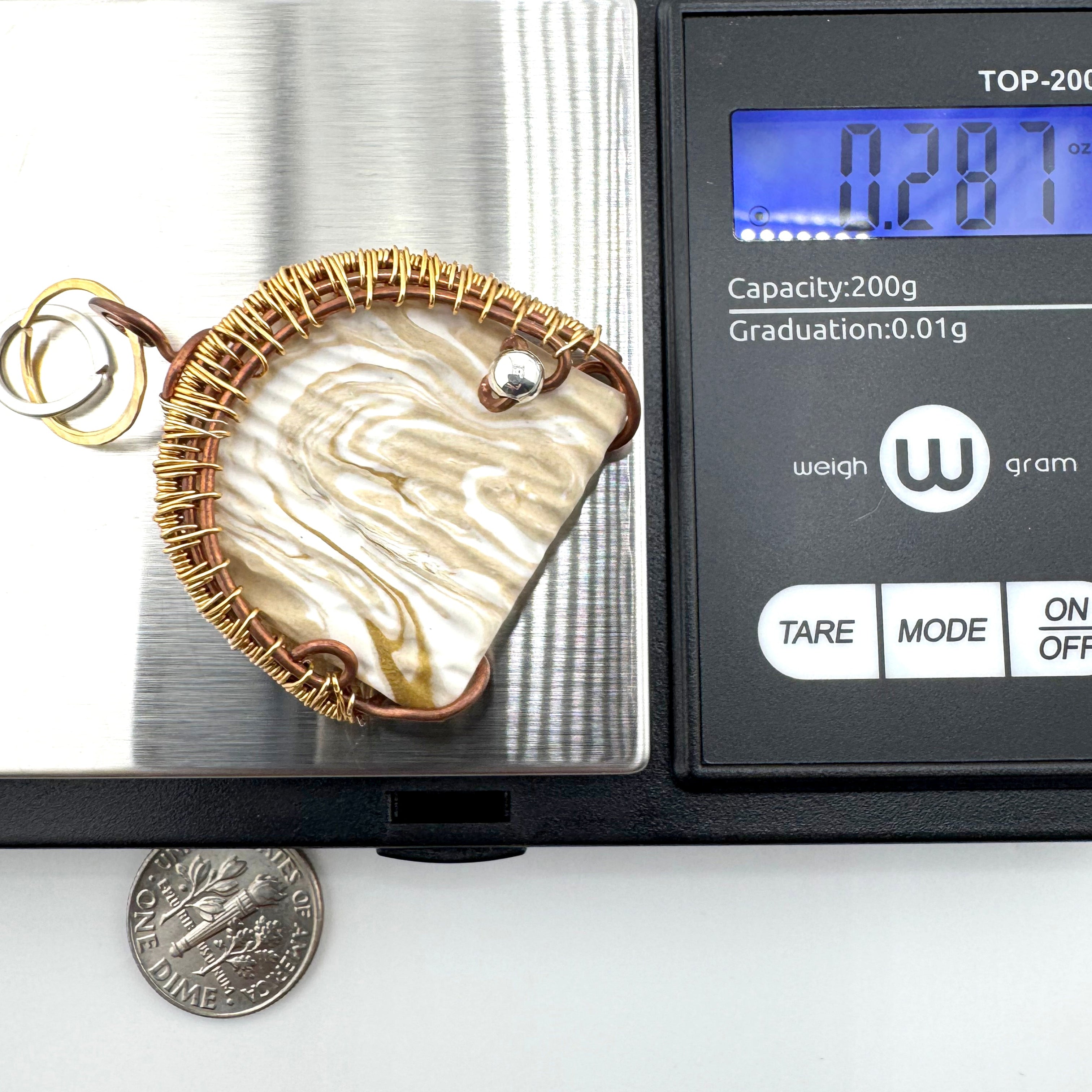 A small clay pendant with a gold-toned woven border and two rings, shown on a digital scale next to a dime for size reference.