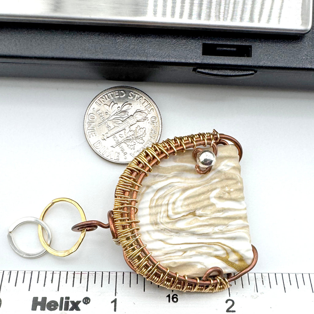 A handmade pendant with a textured cream and white marbled clay inlay, wrapped in gold wire, shown next to a dime and a Helix brand ruler for scale on a white surface.