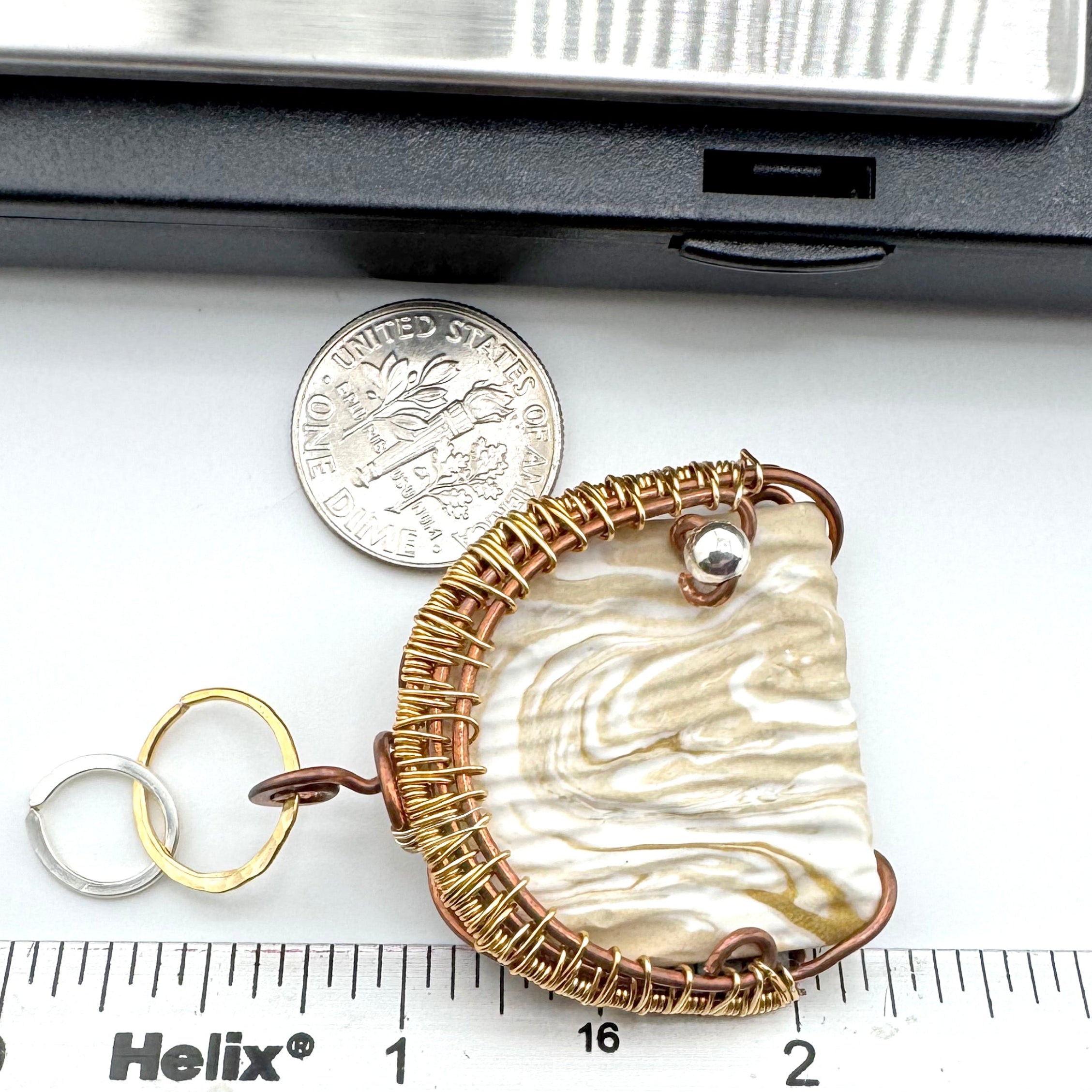A handmade pendant with a textured cream and white marbled clay inlay, wrapped in gold wire, shown next to a dime and a Helix brand ruler for scale on a white surface.