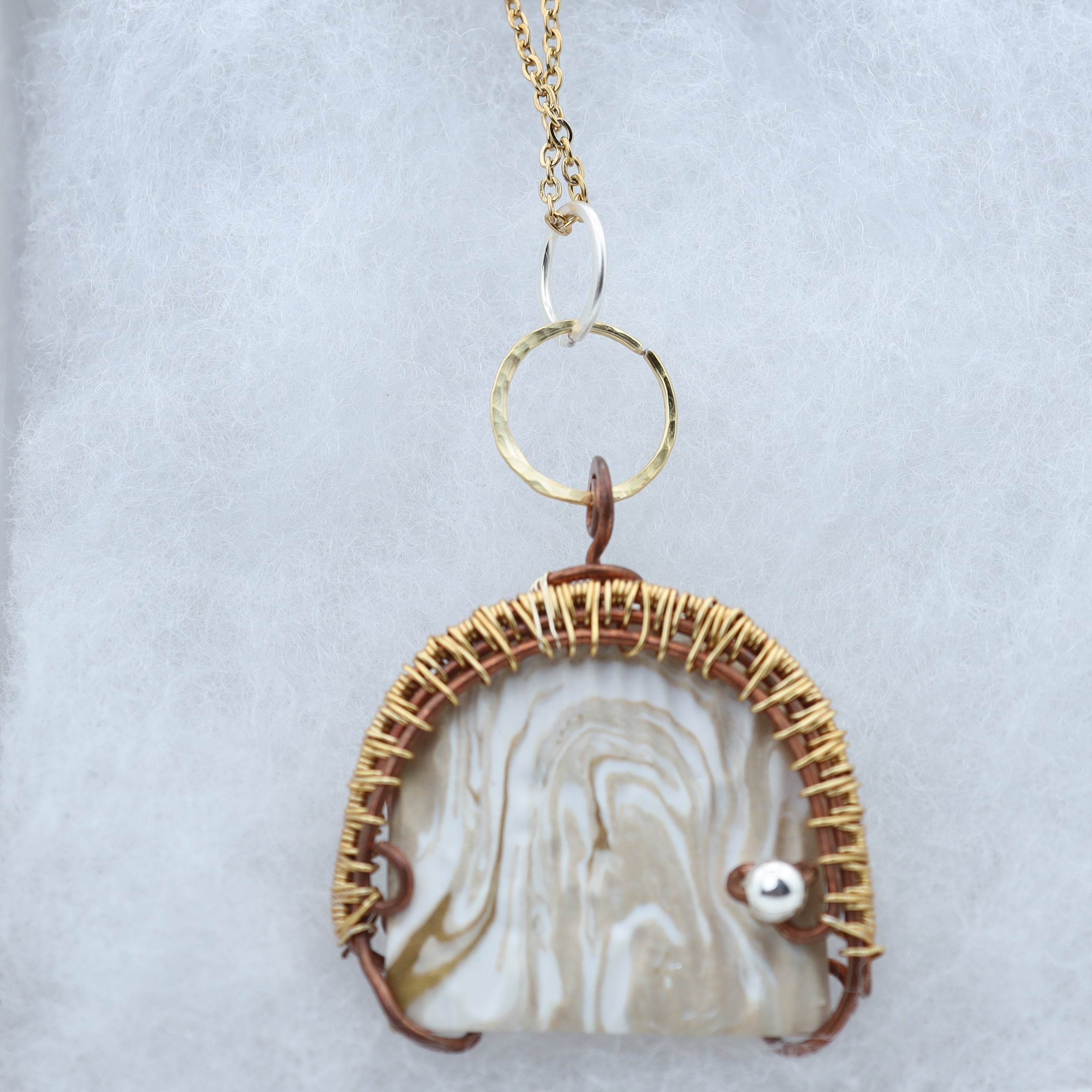 A handmade pendant necklace featuring a marbled cream and white clay focal piece set in a gold wire frame, with a small silver accent and a gold ring connector, displayed on a light gray textured surface.