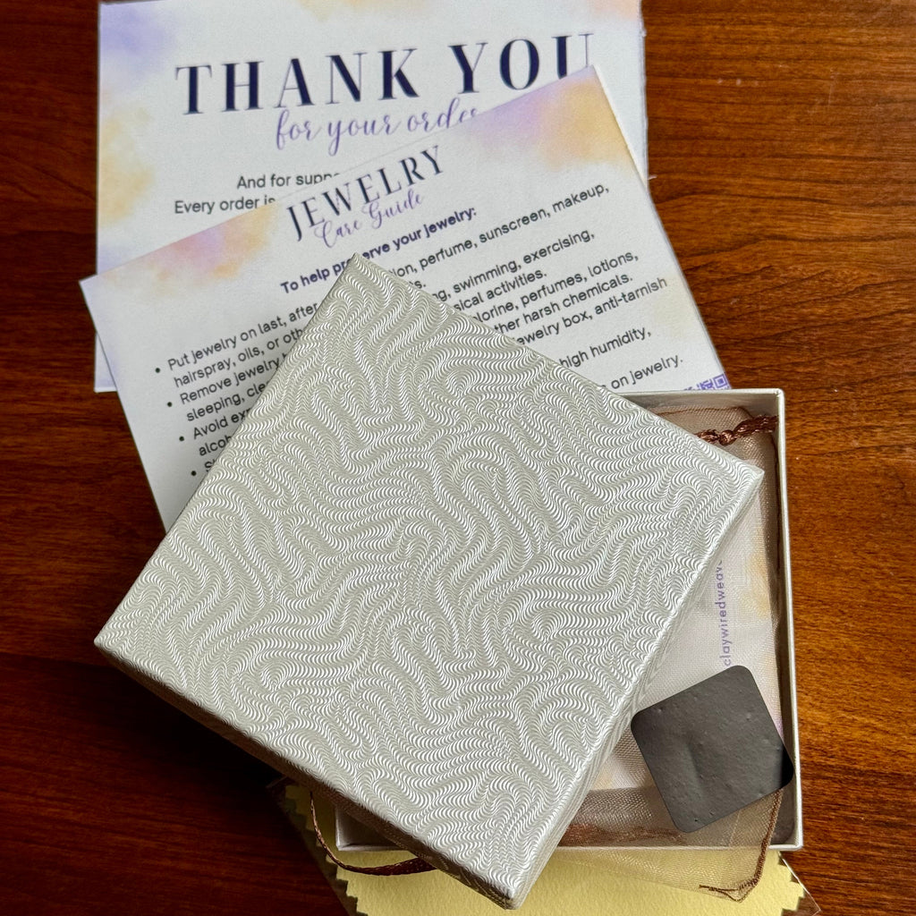 A jewelry gift set on a wooden table, featuring a square white patterned gift box, a gold-colored jewelry bag in the interior, a black, tarnish resistant tag, and a thank you/jewelry care card.