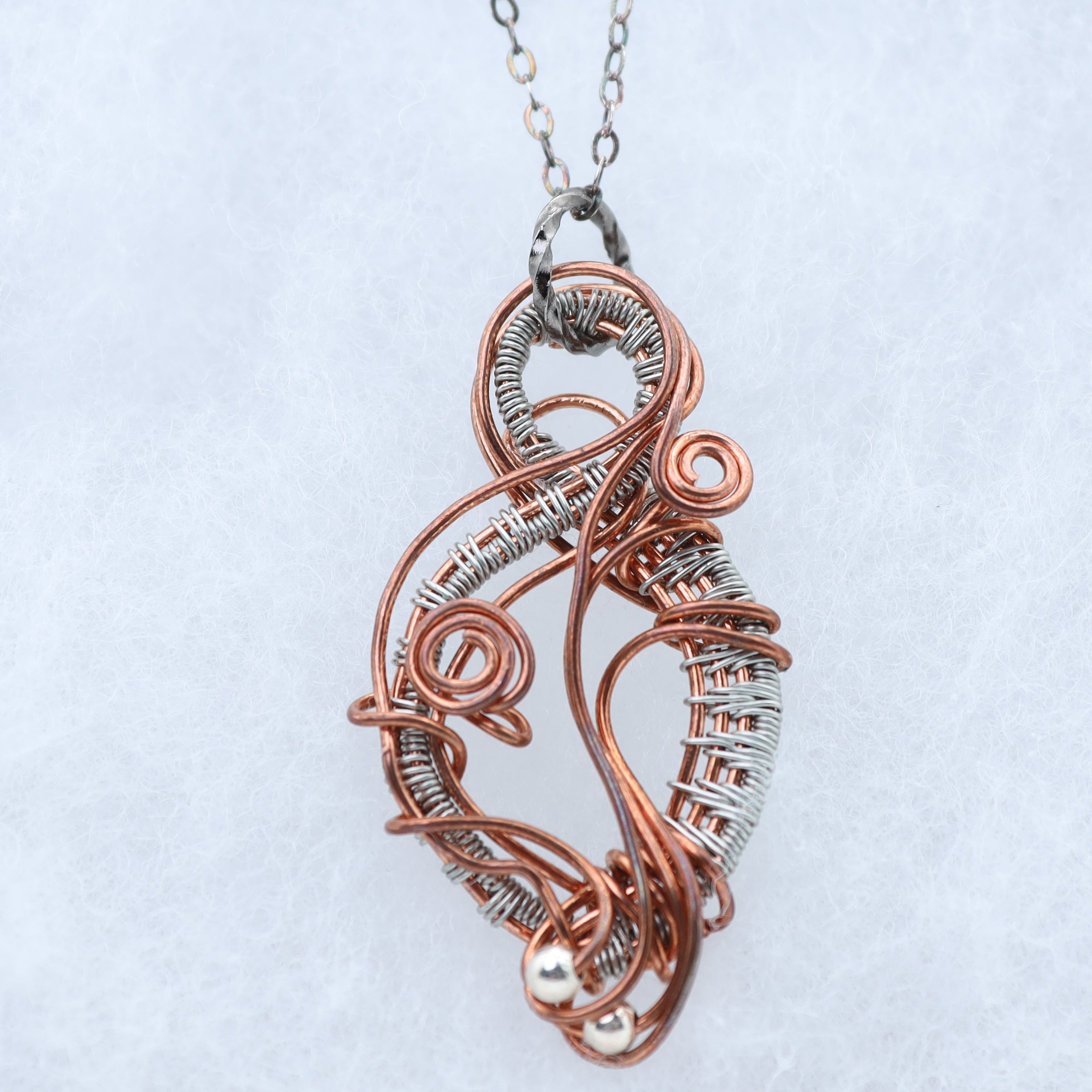 A handmade wire-wrapped pendant necklace featuring copper and silver-toned metal coils, suspended from a silver chain against a light gray textured background.
