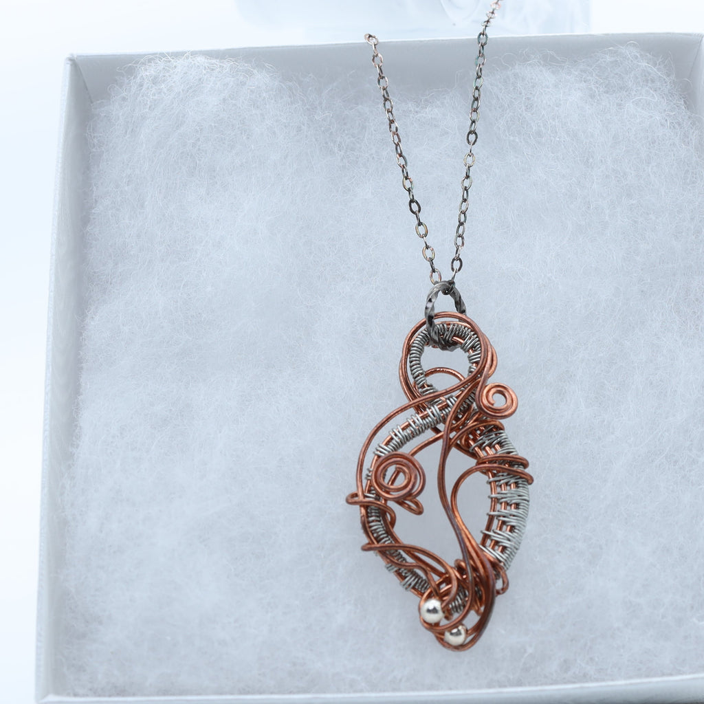 A copper and silver wire-wrapped pendant necklace displayed on a white textured jewelry box.