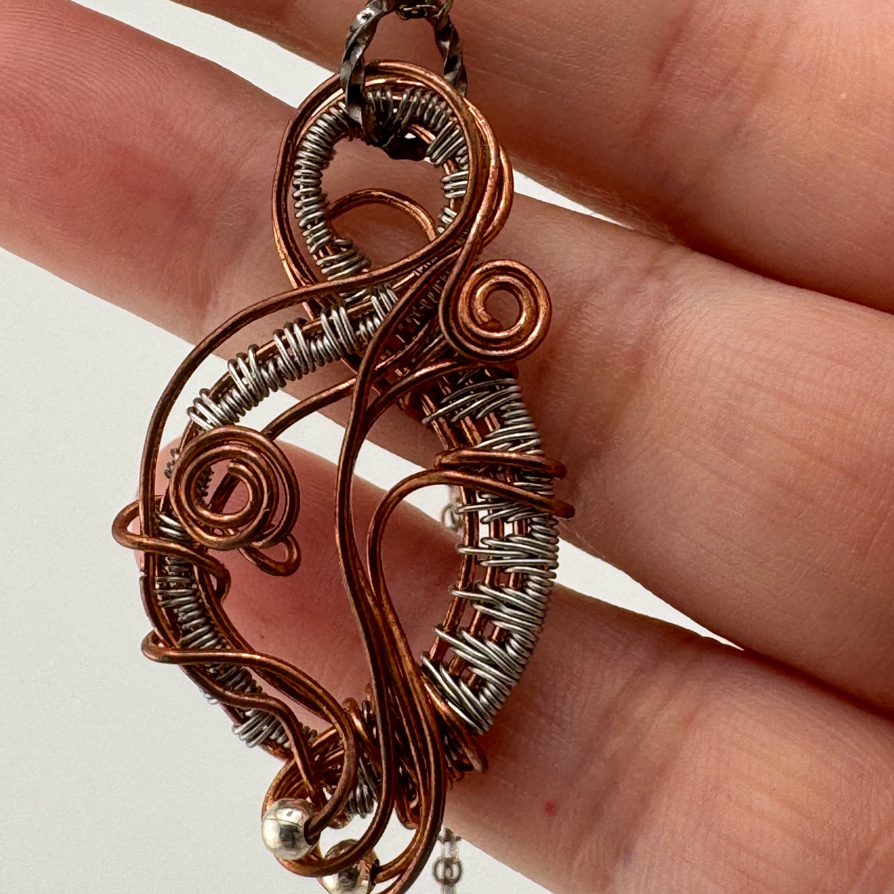 A close-up of a hand holding a copper and silver wire-wrapped pendant necklace against a plain light background.